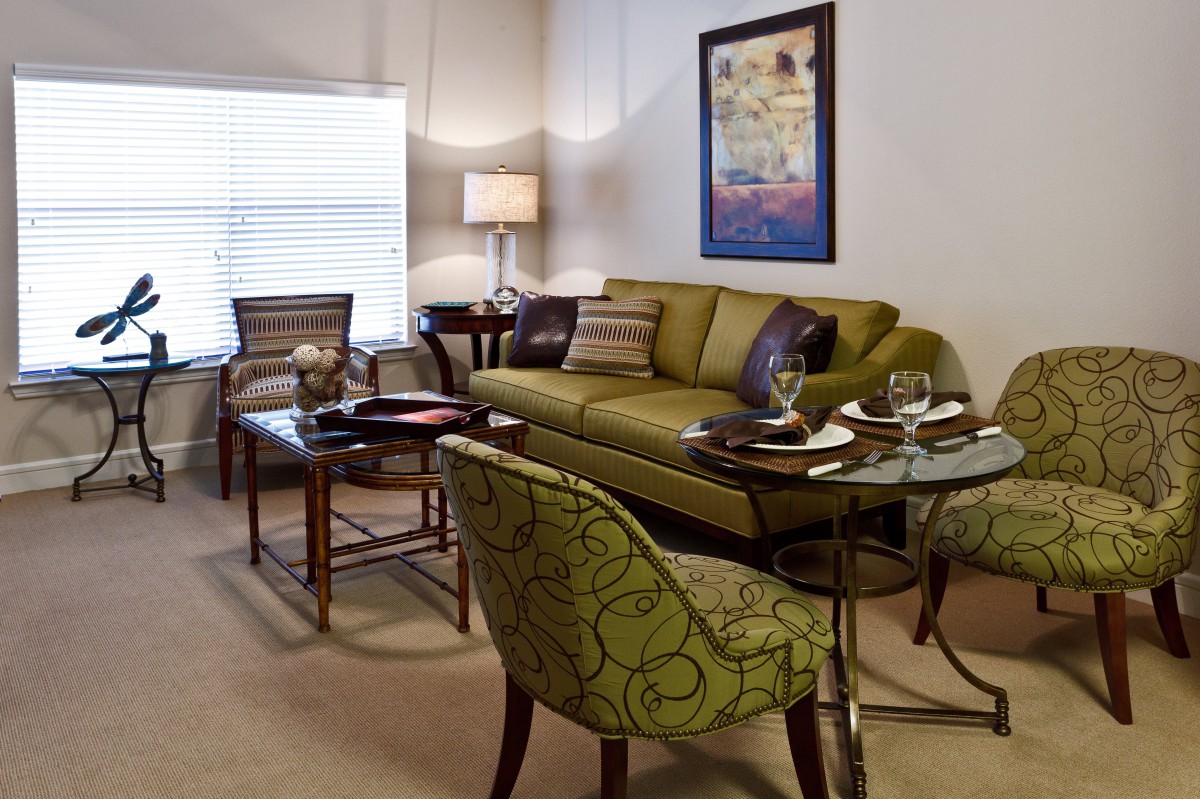 A cozy and elegantly furnished living room with a green sofa, patterned chairs, glass tables, and decorative artwork on the wall.