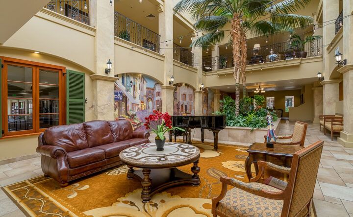 A spacious and elegant lobby featuring a leather sofa, a grand piano, lush indoor plants, and ornate decor.