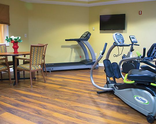 A small gym with exercise equipment including a treadmill and elliptical machine, featuring a wooden floor and a television.