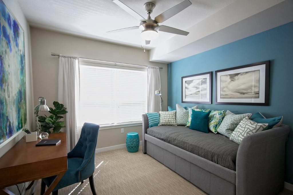 A cozy bedroom with a daybed featuring blue accents and decorative pillows, complemented by a desk and ceiling fan.