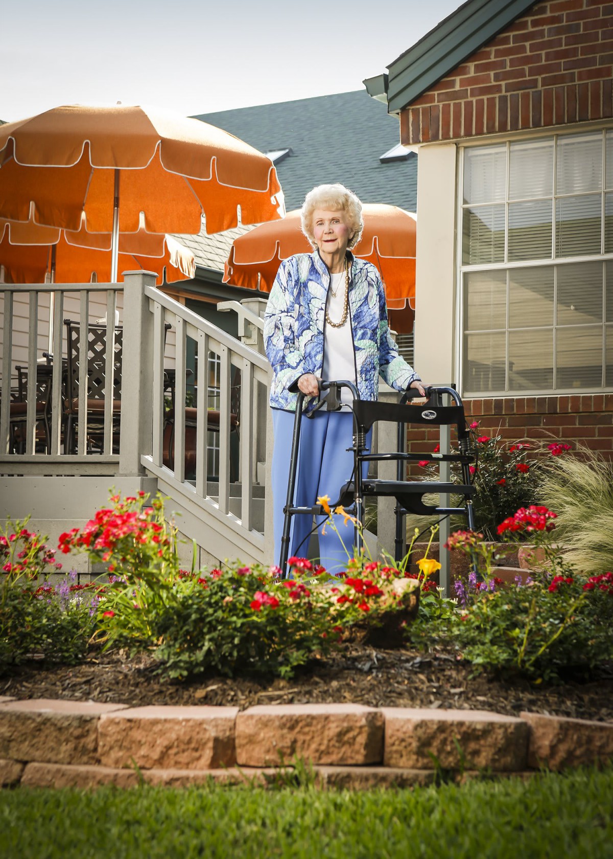 An elderly woman standing with a walker in a garden outside a residential area.