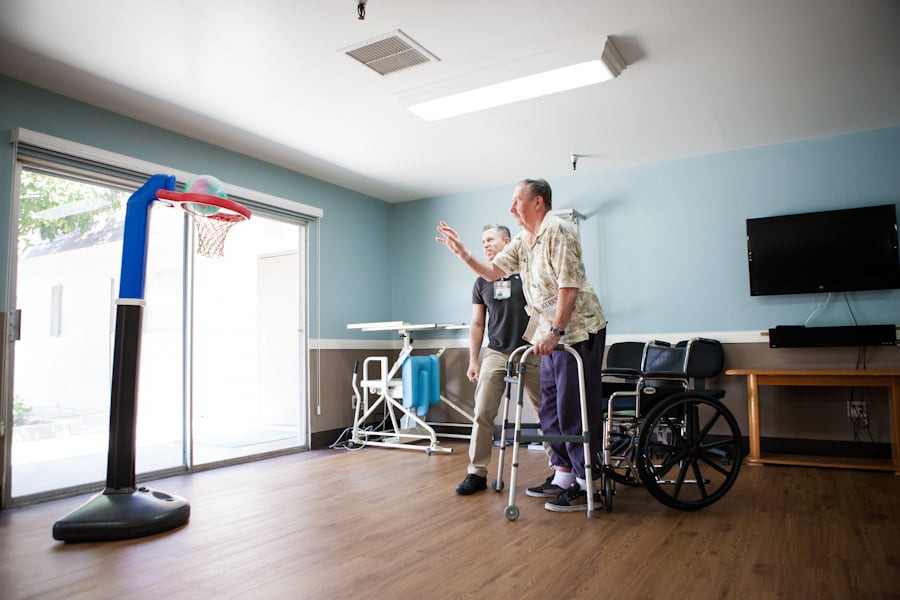 A senior man engages in a therapeutic basketball activity assisted by a caregiver.