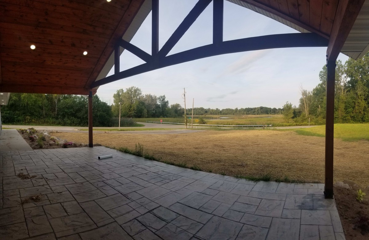 A wide view from a covered patio looking out onto an open field with trees and a clear sky.