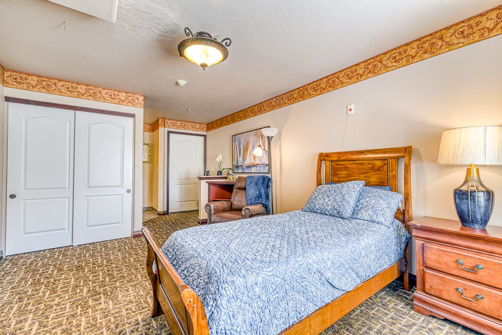 A cozy bedroom with a single wooden bed and blue bedding, featuring carpet flooring and a lamp beside a mirror.