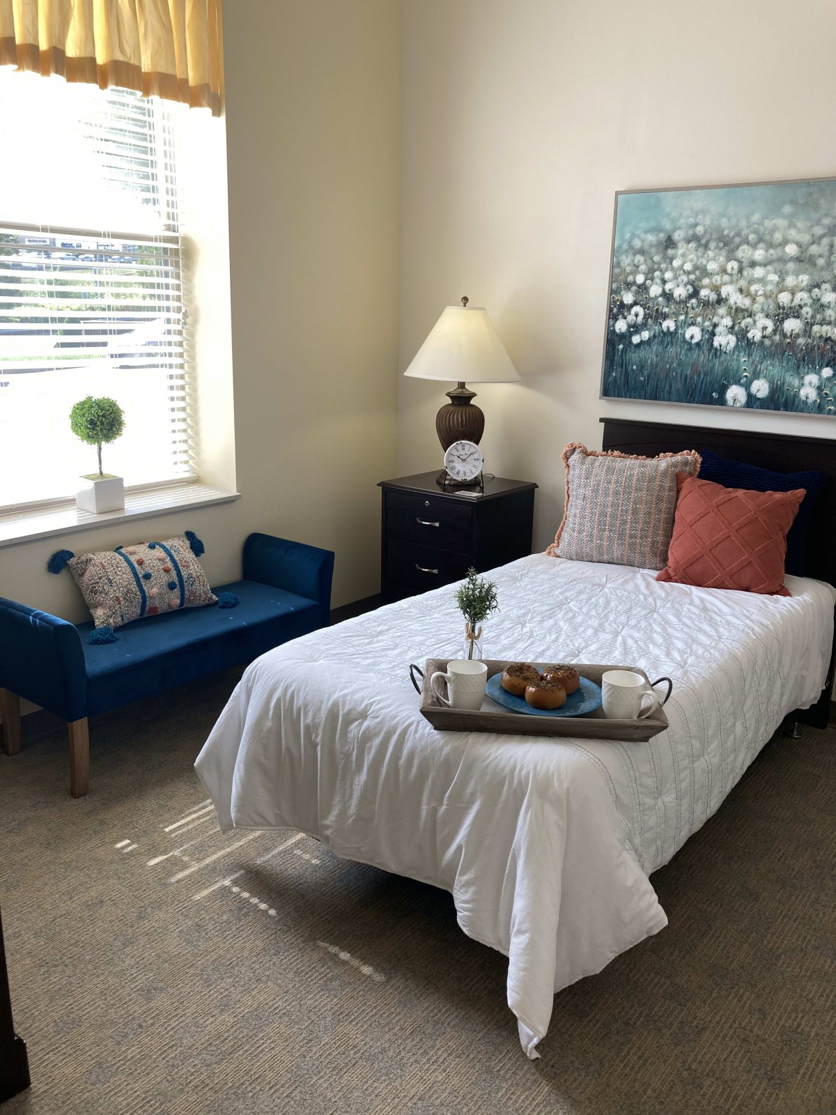 A cozy bedroom featuring a single bed with white linens, a blue bench near the window, and decorative pillows. A lamp and artwork add to the room's ambiance.
