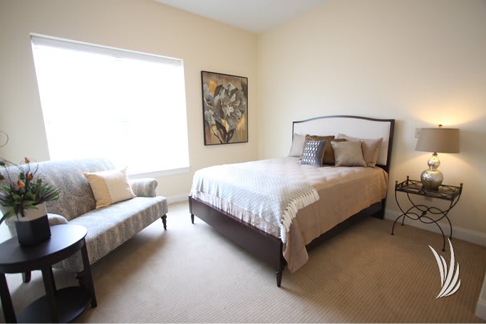 A cozy bedroom with neutral tones, featuring a bed, chair, and wall artwork.