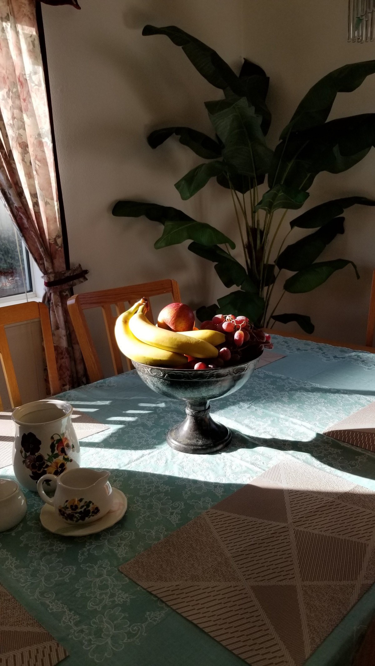 A fruit basket filled with bananas, apples, and grapes sits on a sunlit dining table.