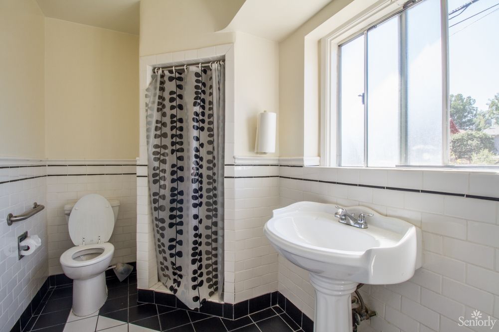 A clean, bright bathroom with a toilet, pedestal sink, and a shower with a patterned curtain.