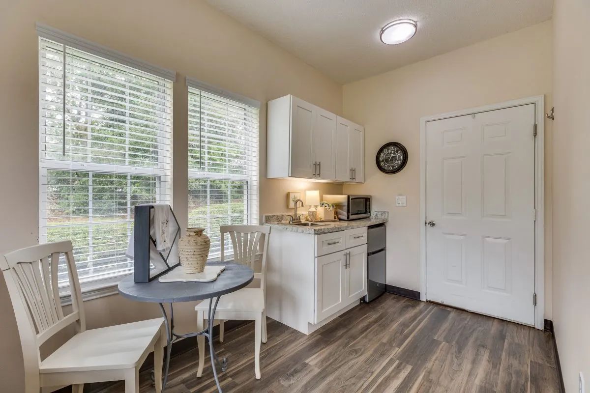 A compact kitchen area with a small dining table and chairs, featuring white cabinets, a microwave, and large windows.