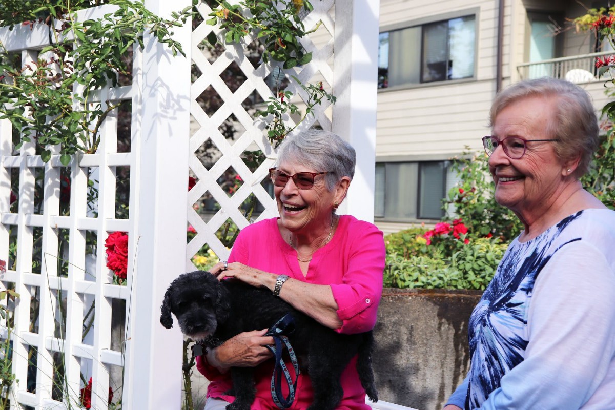 Two smiling women holding a small black dog in a garden setting.