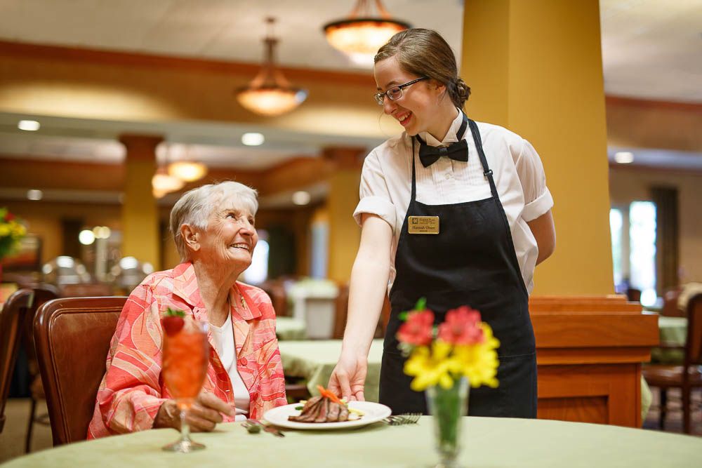 A waitress serves a meal to a smiling senior woman at a restaurant.