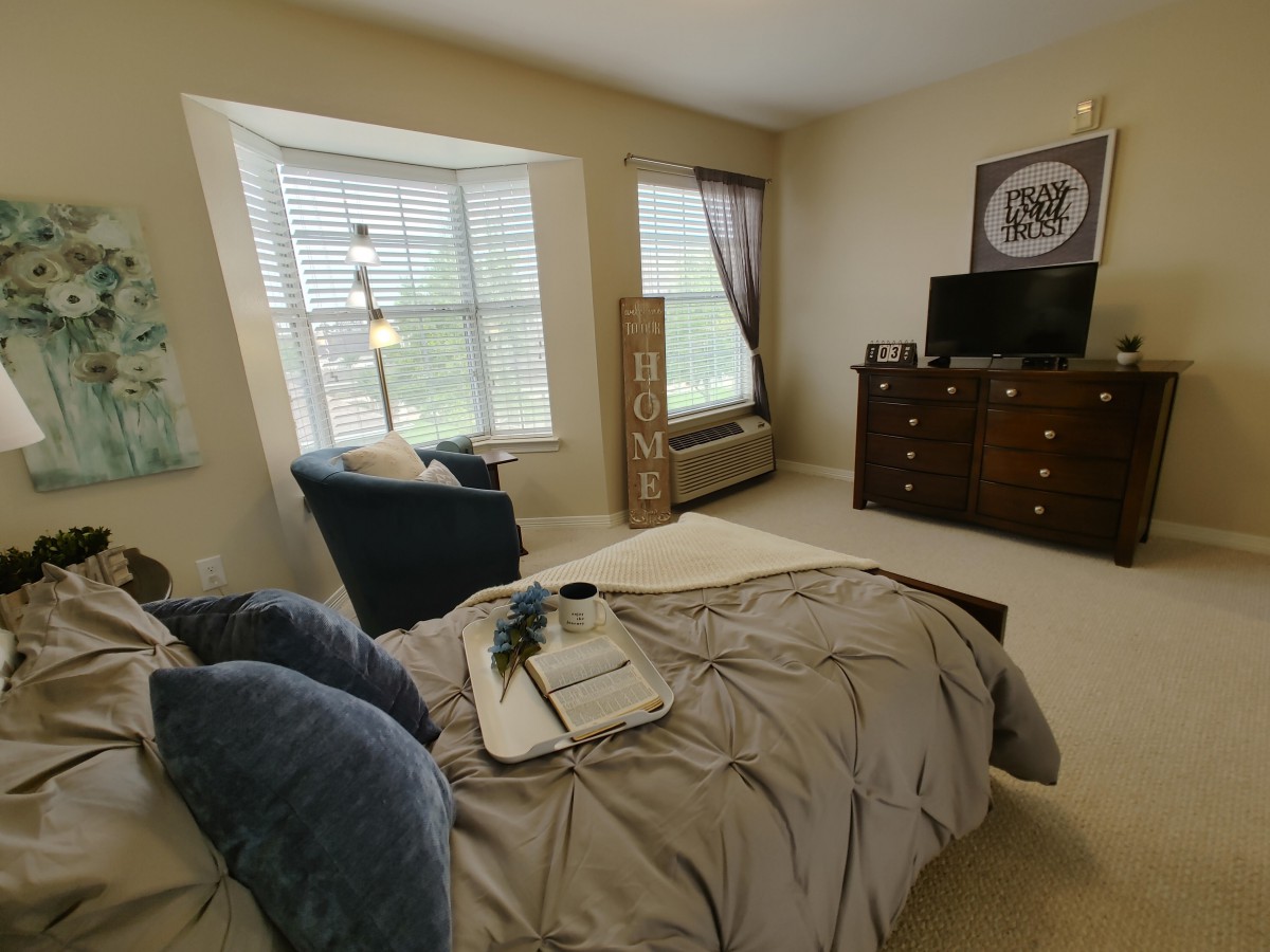 A cozy bedroom with a neatly made bed, featuring blue accent pillows and a tray. The room includes a TV on a dresser and a comfortable chair by the window.