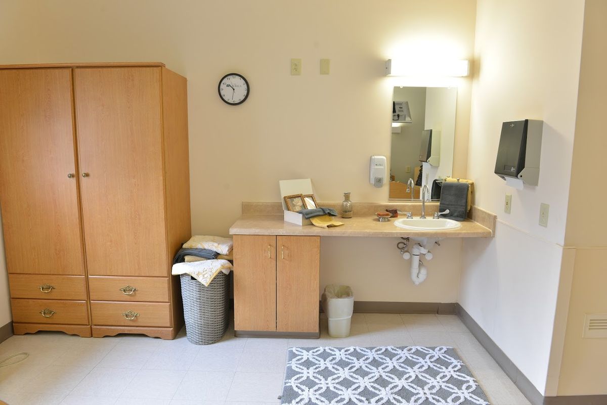 A simple room interior with a wooden dresser, a sink with a mirror above it, a clock on the wall, and a patterned rug on the floor.