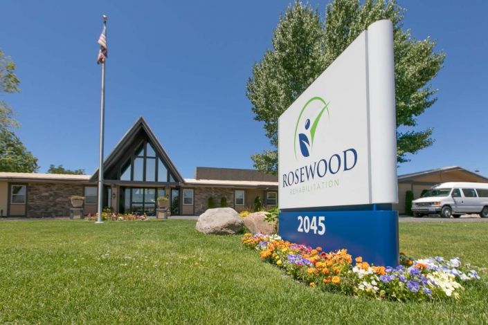 A building with a sign in front displaying 'ROSEWOOD' and colorful flowerbeds under a clear blue sky.