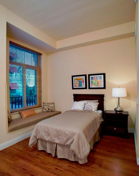 A cozy bedroom with a single bed and window seat, featuring a nightstand with a lamp and colorful artwork on the wall.