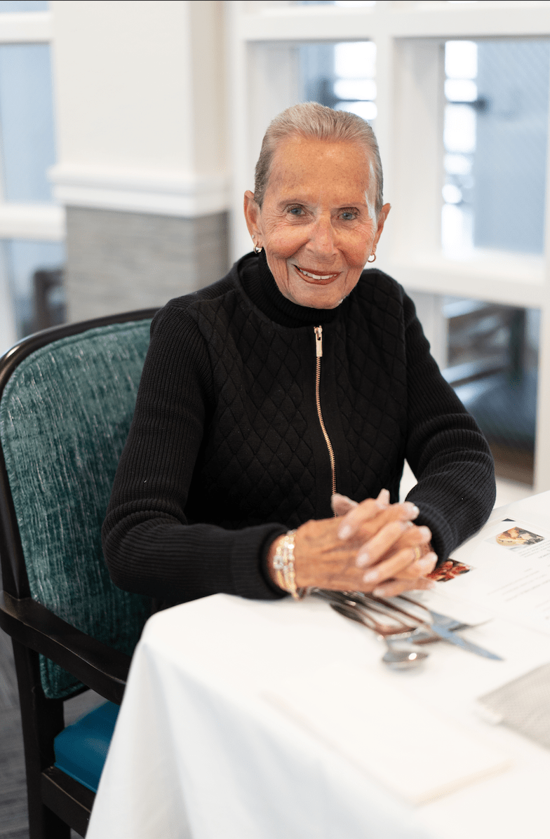 An elderly woman sitting at a table indoors, smiling.
