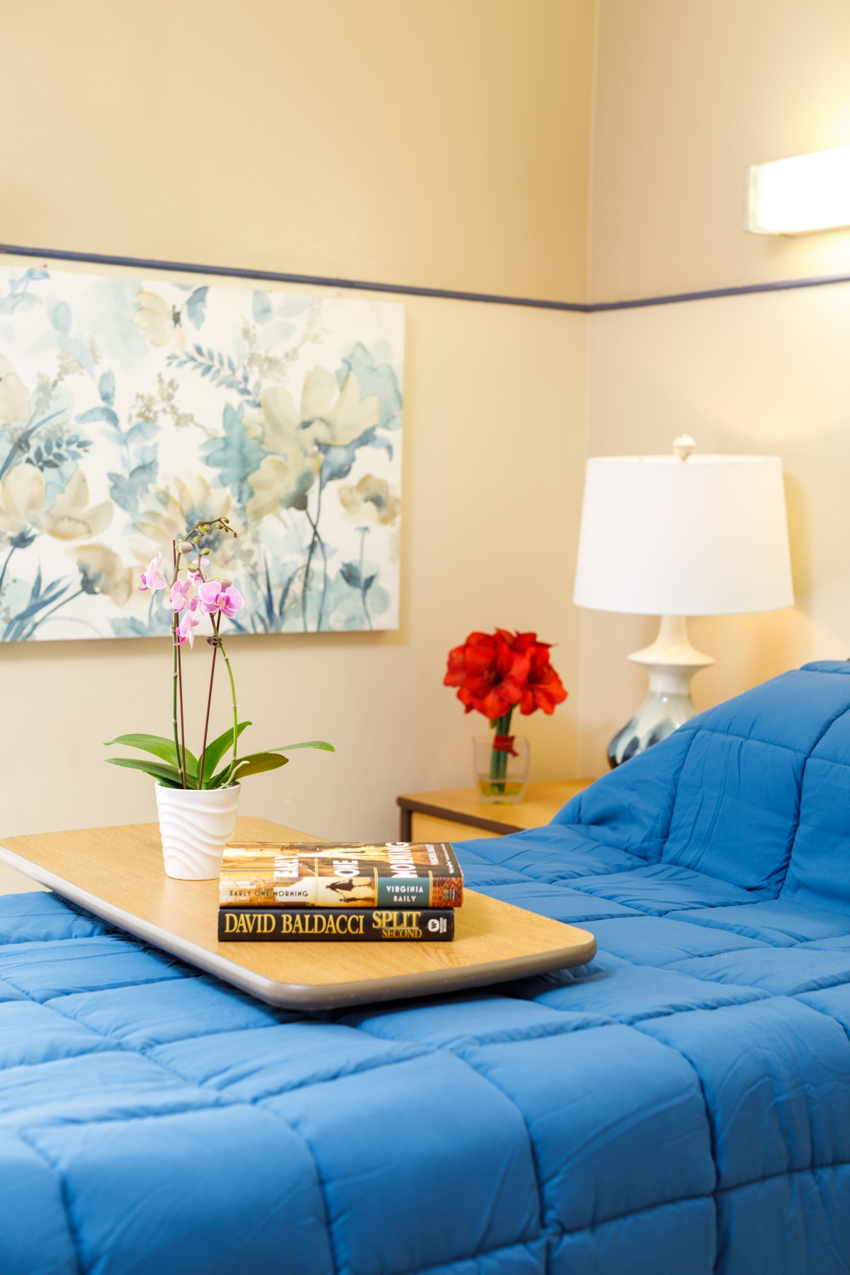 A cozy bedroom setup featuring blue bedding, a side lamp, wall art, and a tray with books and an orchid plant.