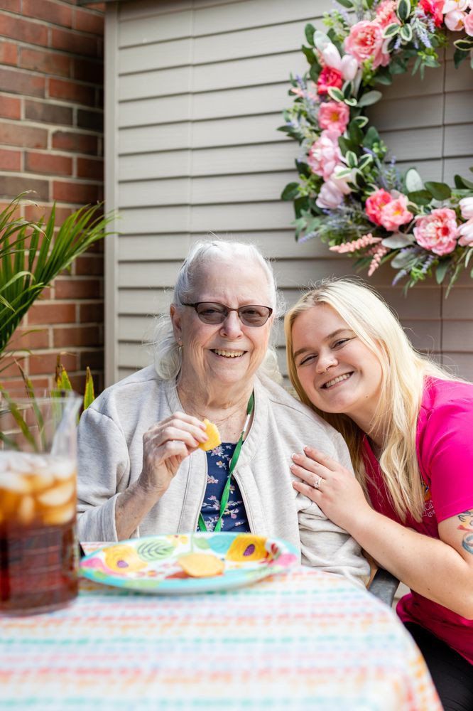 An elderly woman and a younger woman smiling together outdoors, enjoying a snack by a floral arrangement.