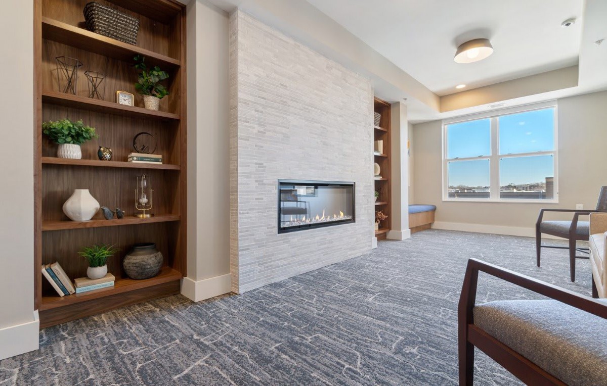 A modern living room with a white stone fireplace, built-in bookshelves, and large window.