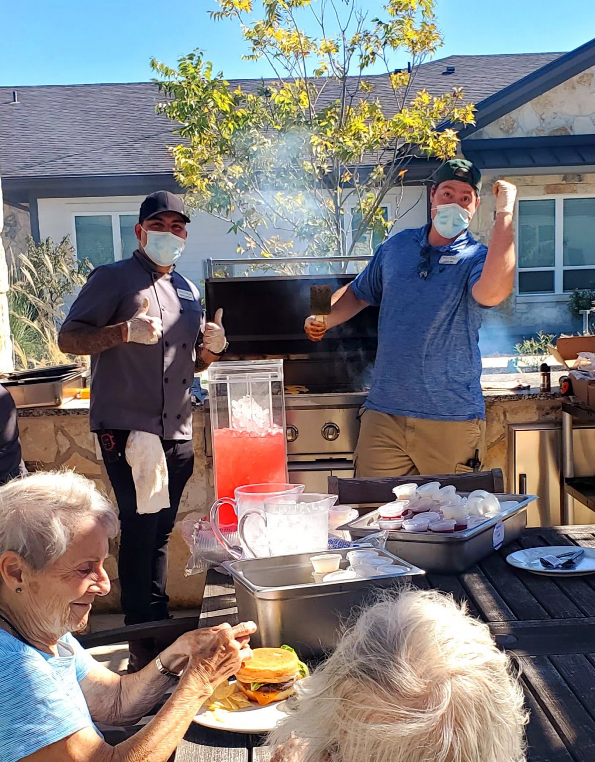 Two people are cooking at an outdoor barbecue, wearing masks, as others gather around.