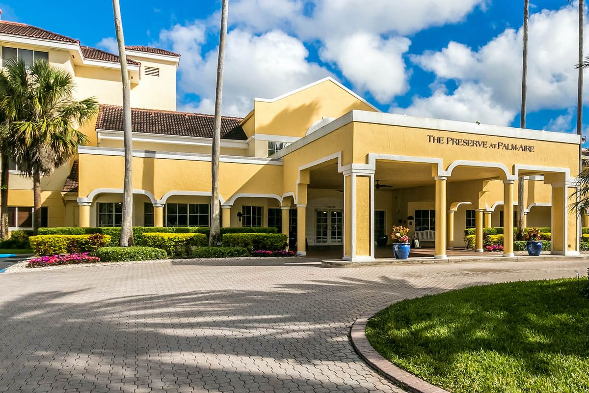 A yellow hotel building with a paved driveway, surrounded by palm trees under a cloudy sky.