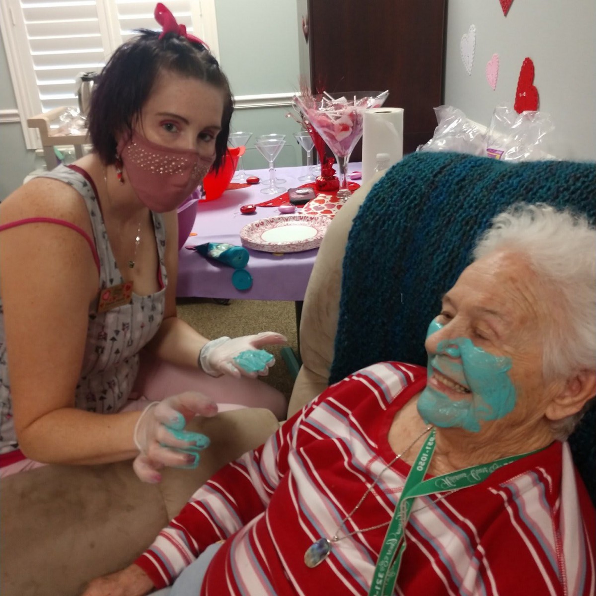 An elderly woman enjoys a spa day at home with a facial mask, assisted by her caregiver.
