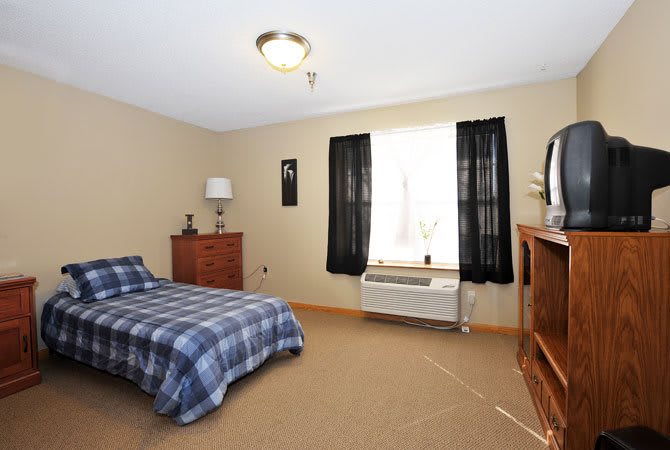 A simple bedroom featuring a single bed, dresser, and television with natural light from a window.