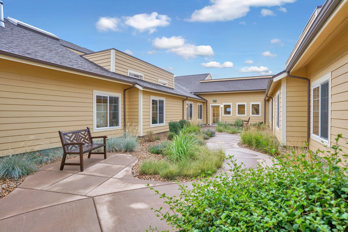 A peaceful courtyard with a bench surrounded by greenery and modern building exteriors under a partly cloudy sky.