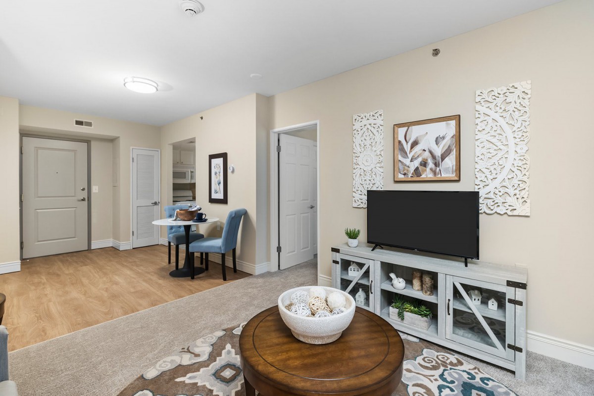 A modern living room with neutral colors, featuring a TV, artwork on the walls, and a small dining area.