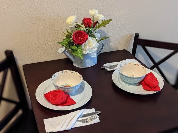A cozy dining setup with two place settings, featuring elegant bowls, red napkins, and a centerpiece of red and white flowers.