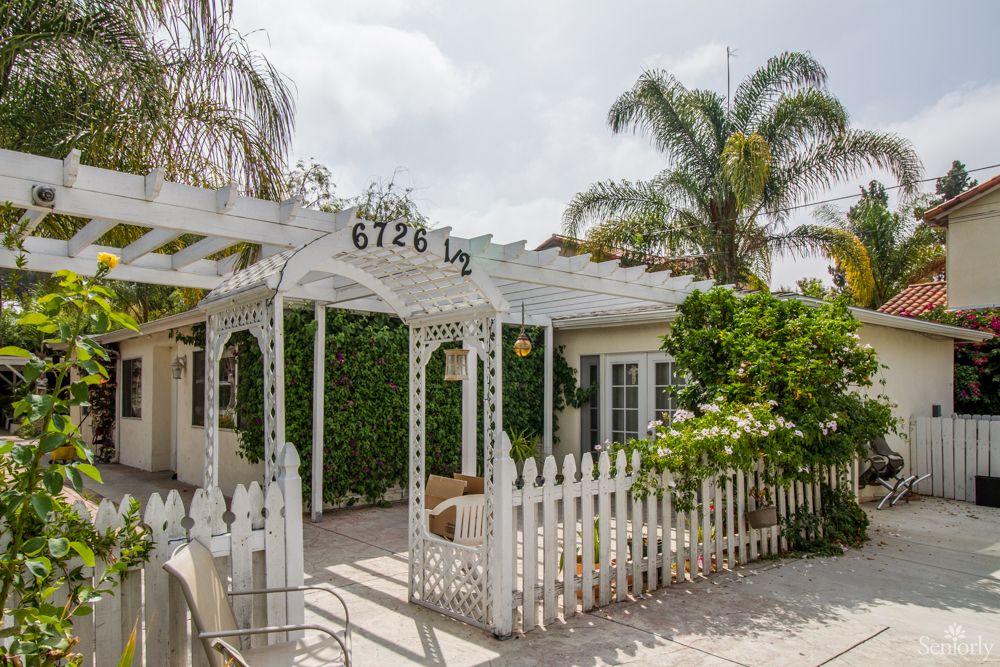 Charming house with a white picket fence and pergola, surrounded by lush greenery and palm trees.