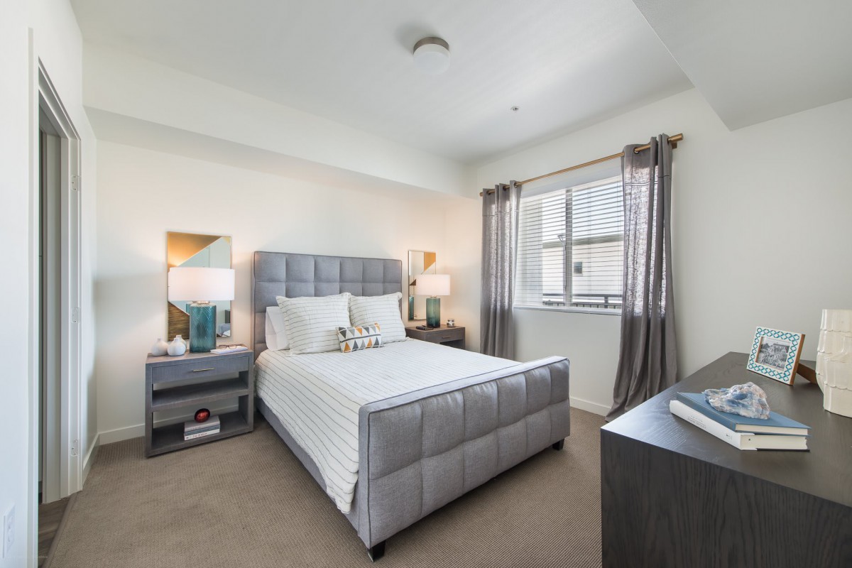 A modern bedroom with a sleek design, featuring a gray padded bed, side tables with lamps, and a large window allowing natural light.