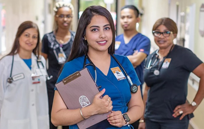 A nurse stands in a hospital corridor with her colleagues, holding a clipboard.