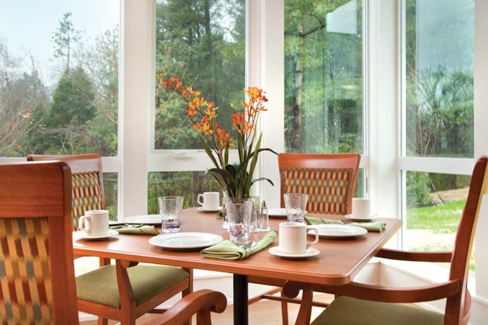 A dining table set with plates, cups, and a flower vase, surrounded by wooden chairs in a bright room with large windows.