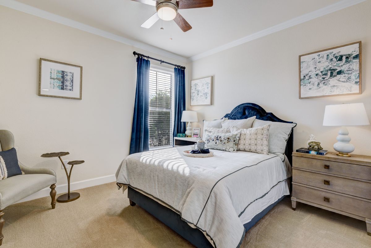 A cozy bedroom with a neatly made bed featuring blue accents, a lamp on the nightstand, and natural light coming through the window.