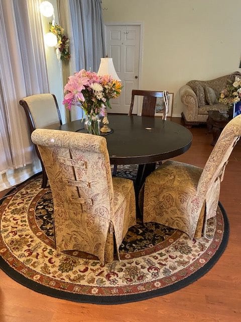 A cozy dining room featuring a round table with upholstered chairs, floral centerpiece, and a decorative carpet.