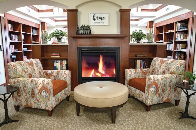 A cozy living room with a fireplace, two patterned chairs, and a round ottoman.