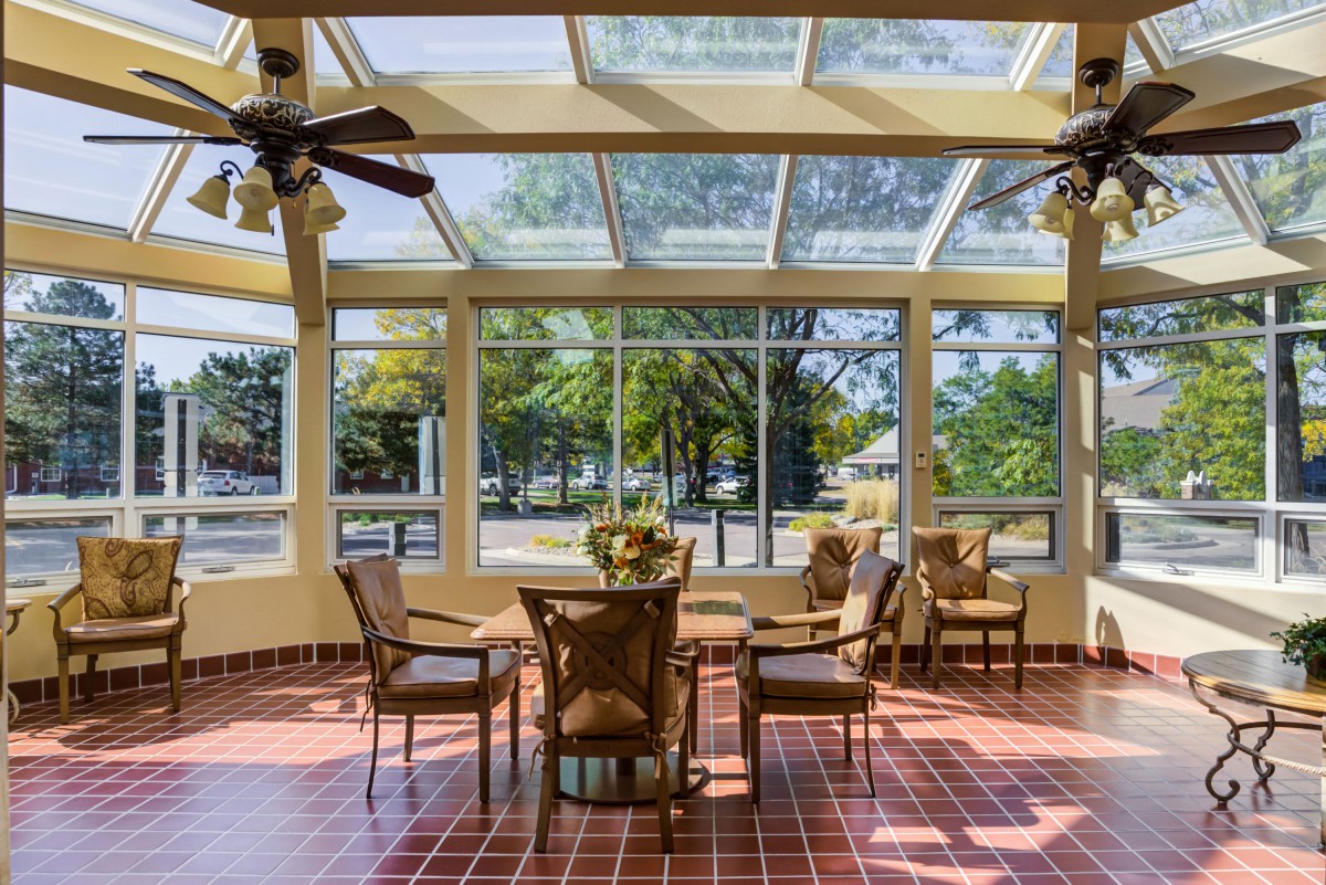A sunlit room with large windows, featuring a table and chairs, surrounded by greenery.
