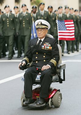 A military veteran in formal uniform participates in a parade while seated in a motorized wheelchair, holding an American flag.