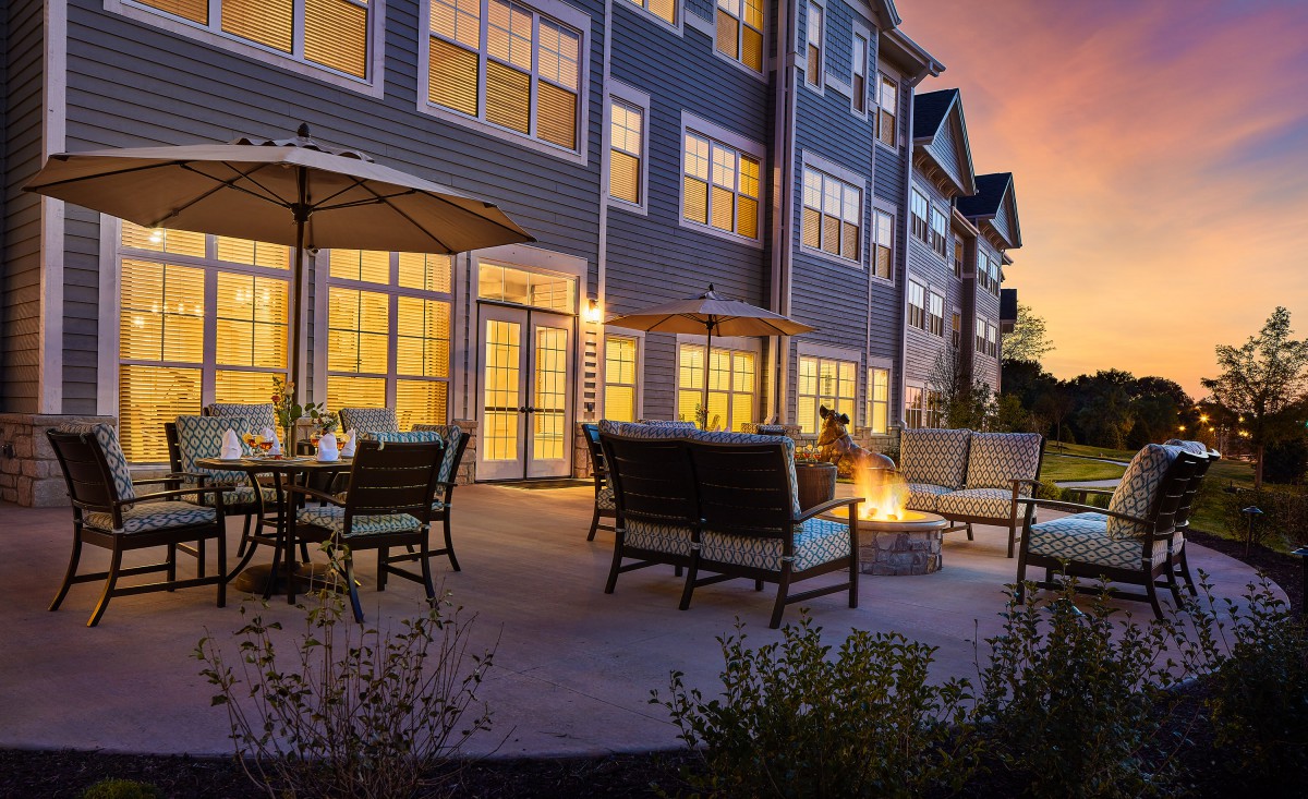 A cozy outdoor patio with comfortable seating and a fire pit, set against an apartment building during sunset.