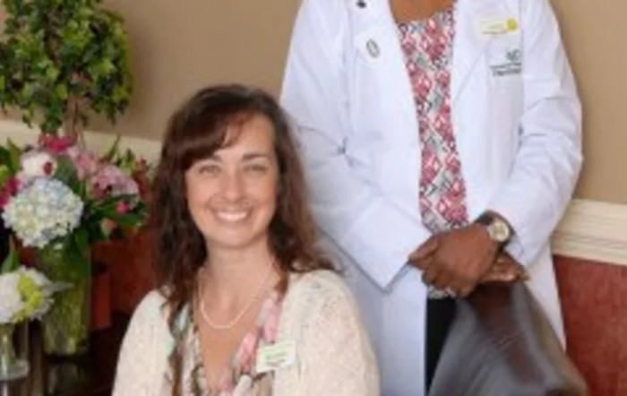 A professional setting with two individuals, one seated and smiling, the other standing in a lab coat.