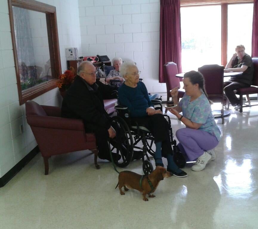 A caregiver interacts with two elderly individuals in a nursing home setting, accompanied by a small dog.