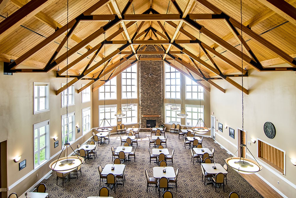 A spacious dining hall with wooden beams and large windows, featuring neatly arranged tables and chairs.
