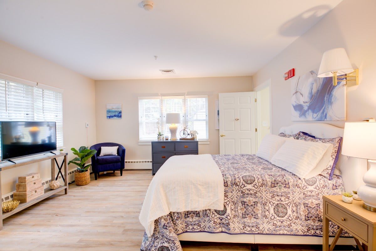 A bright and spacious bedroom featuring a patterned bedspread, an armchair with a plant beside it, and a large window allowing natural light to fill the room.