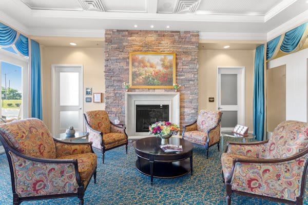 A cozy living room with a fireplace, surrounded by elegant chairs and a central coffee table adorned with a floral arrangement. The walls feature a serene painting above the mantel.