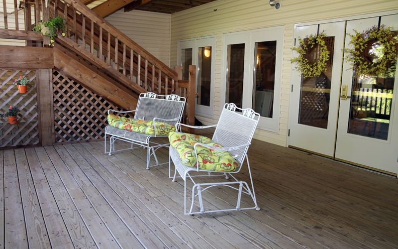 A cozy porch area featuring two metal chairs with floral cushions on a wooden deck, alongside a welcoming door adorned with a wreath.