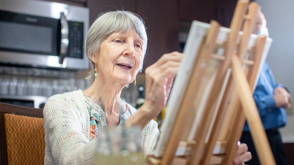 An elderly woman painting at an easel, engaged in creative activity indoors.