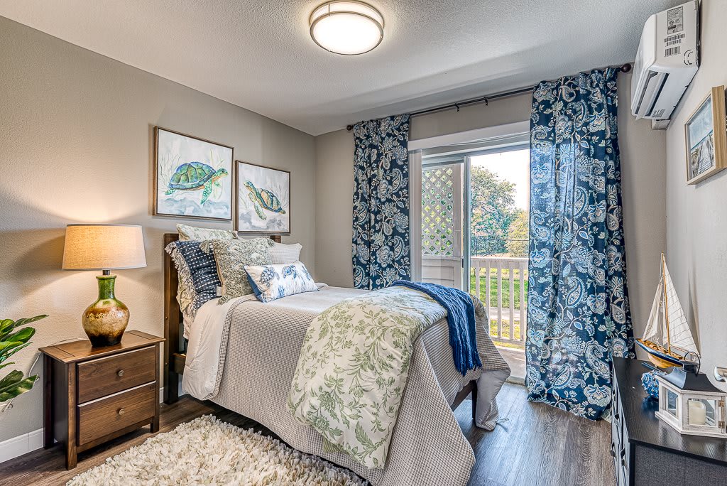 A cozy bedroom with blue and beige decor, featuring a single bed, artwork on the walls, and a window with floral curtains.