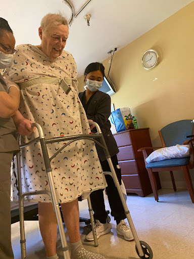 Two caregivers assist an elderly person using a walker in a healthcare setting.