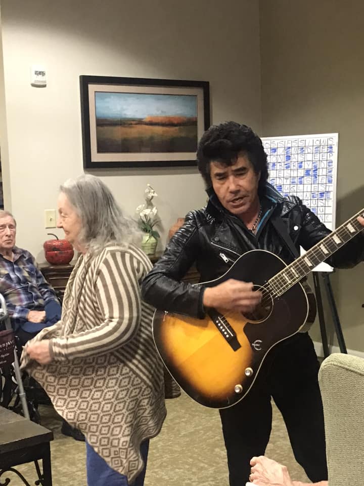 A musician playing guitar indoors with an audience of elderly people.